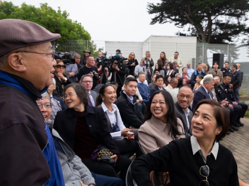 A man stands and talks to seated people at an outdoor event with a crowd, photographers, and trees in the background.