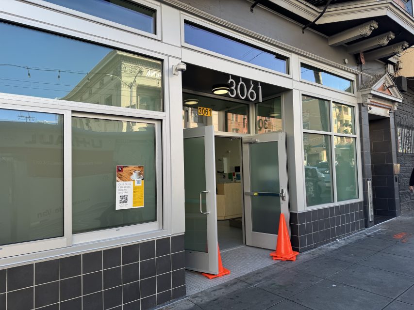 A modern storefront at 3061 with large windows, open glass doors, two orange cones at the entrance, and a poster with a QR code on the left window.