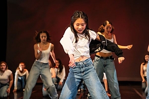 A group of young people in casual clothing perform a dance routine on stage, with one dancer in front wearing a white shirt and blue jeans.