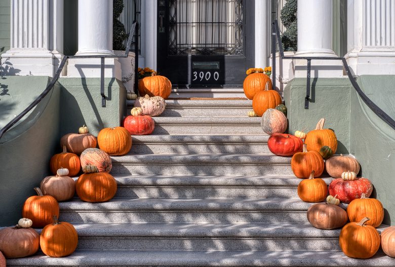 A variety of pumpkins and gourds decorate the steps leading to a black door numbered 3900, flanked by white columns and green railings.