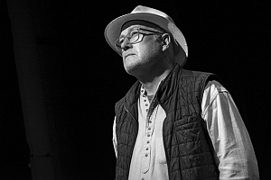 A man wearing glasses, a light-colored hat, and a buttoned shirt with a quilted vest stands against a dark background. The image is in black and white.