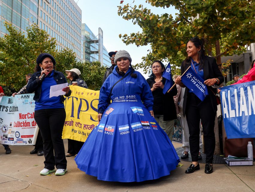 A group of women stand outside at a rally holding signs and banners advocating for TPS, with one woman wearing a blue skirt decorated with country names.