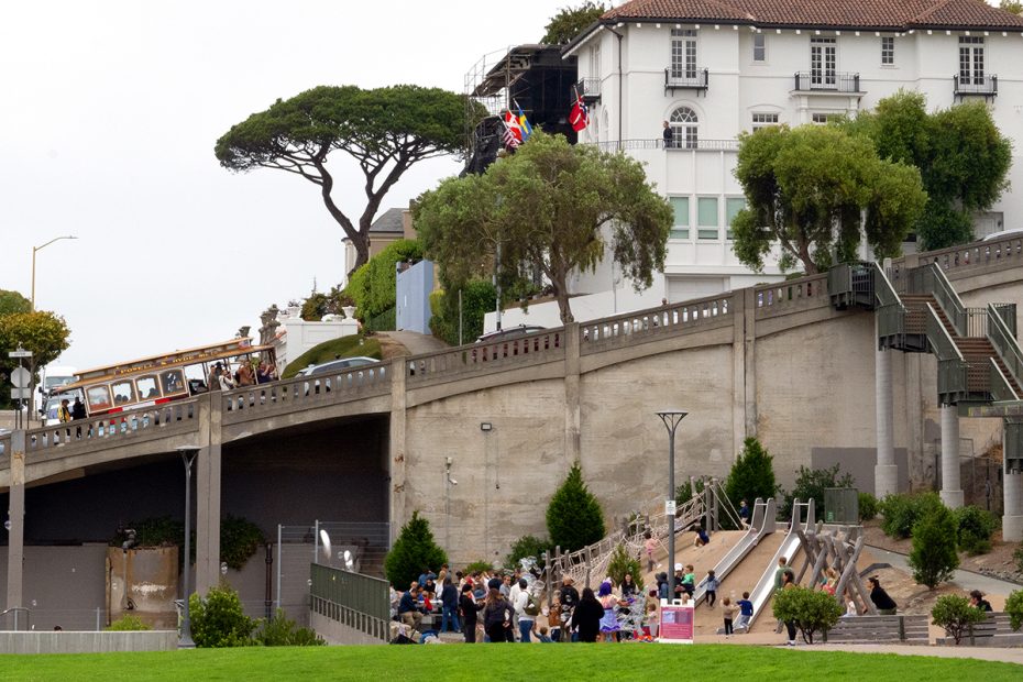 A group of people gathers at the bottom of concrete slides in a park, while a cable car ascends a steep street in front of historic buildings and trees.