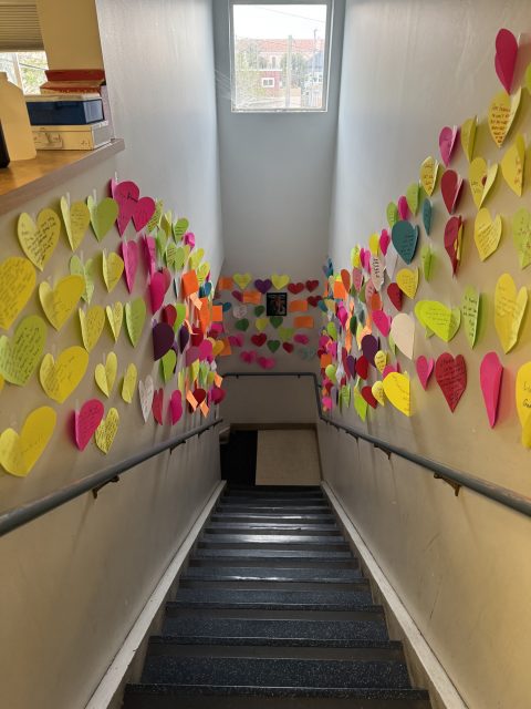 A stairwell with walls covered in colorful paper hearts, each containing handwritten messages, leading down to a landing with a window above.