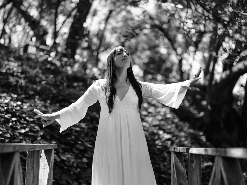 A woman in a long white dress stands on a wooden bridge in a forest, arms outstretched, looking upward. The image is in black and white.
