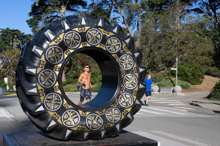 A shirtless man jogs past a large black circular sculpture with gold designs, while another person walks on a crosswalk in a sunny park setting.