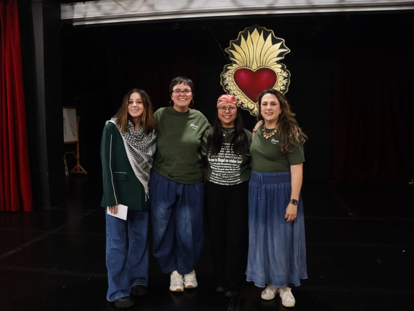 Four women stand together smiling on a stage, with a large heart-shaped artwork in the background. Three wear green tops and jeans or skirts; one wears black.