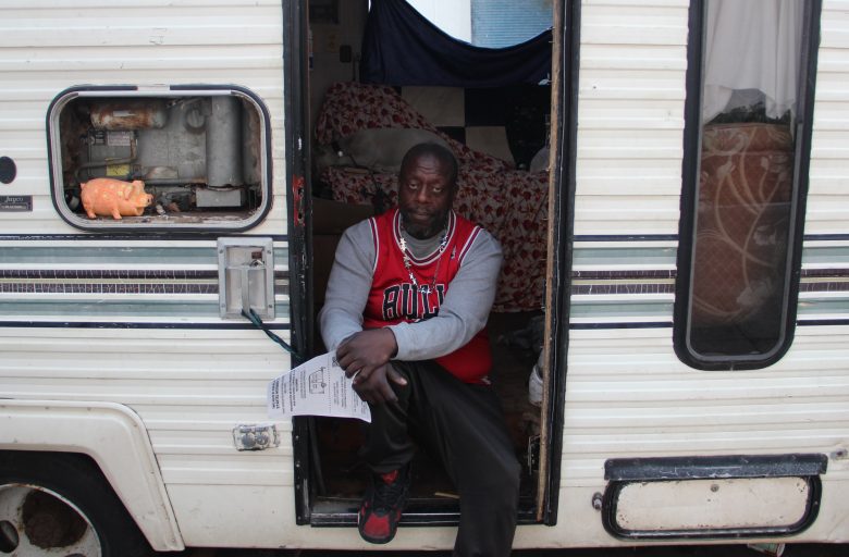 A man in a red sports jersey sits in the doorway of an RV camper van, holding a paper, with a bed and a small shelf visible inside.