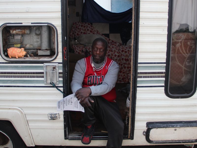A man in a red sports jersey sits in the doorway of an RV camper van, holding a paper, with a bed and a small shelf visible inside.