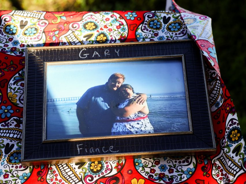 A framed photo labeled "Gary" and "Fiance" shows a couple embracing on a beach, placed on a colorful, patterned cloth with skull designs.