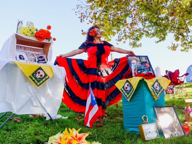 A woman in a red and black dress poses outdoors between two decorated altars, with a Puerto Rican flag and framed photos displayed on the grass.
