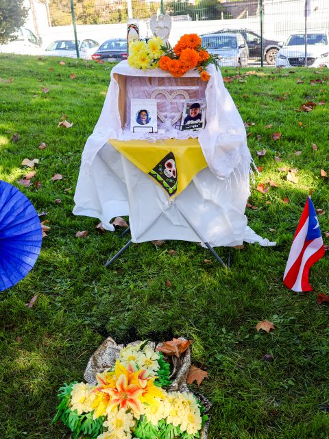 A small memorial altar with photos, flowers, and flags is set up outdoors on grass, surrounded by decorations and fallen leaves.