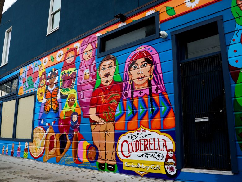 A colorful mural on a building exterior features portraits, pastries, and patterns, with a sign reading "Cinderella Russian Bakery Cafe.