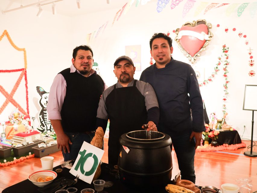 Three men stand behind a food station with a soup pot at an indoor event decorated with colorful papel picado and Day of the Dead motifs.
