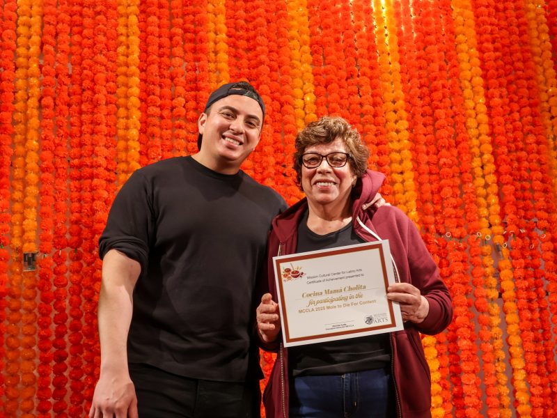Two people stand in front of an orange and yellow marigold backdrop; one holds a certificate recognizing Lorina Maza Chaltlat for her tacos in Ventura County.