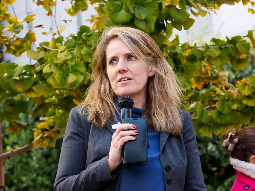 A woman in a blazer holds a microphone and a smartphone, speaking outdoors in front of a leafy tree.