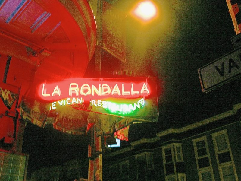 Neon sign for "La Rondalla Mexican Restaurant" glows at night on a street corner, with a streetlight and "Valencia" street sign visible.
