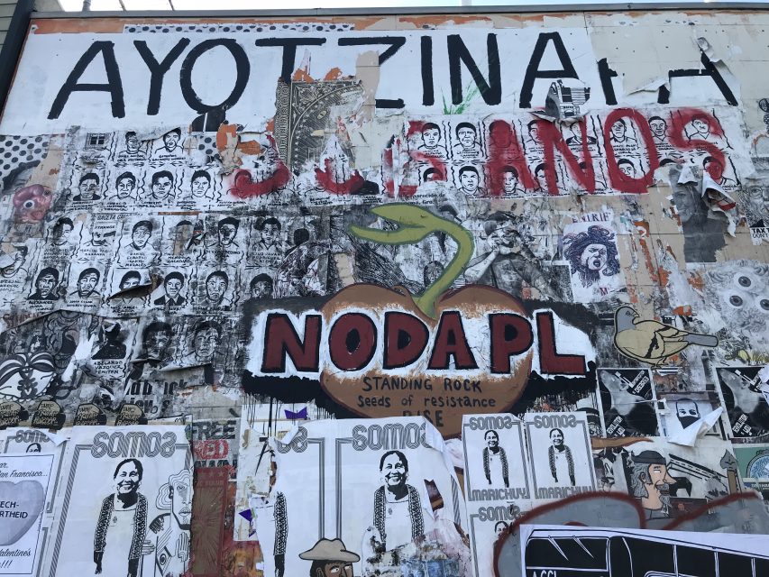 Art wall covered with protest art, posters, and graffiti, including "AYOTZINAPA," "NODAPL," and faces—powerful visuals referencing social justice movements and resistance.