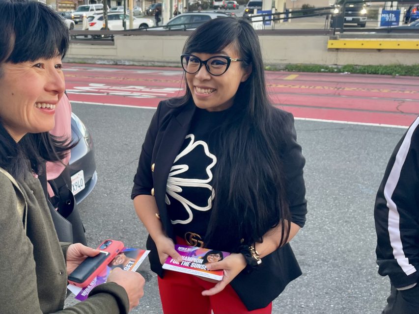 Two women stand on a city street. One woman smiles while holding campaign flyers with images and text. Cars and people are visible in the background.