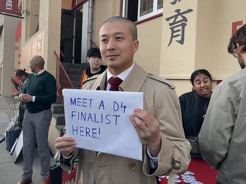 A man in a beige trench coat holds a sign that reads “MEET A D4 FINALIST HERE!” while standing outside near a group of people.