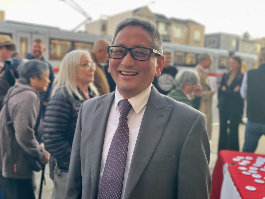 A man in a suit and tie smiles at the camera while standing outdoors among a group of people.