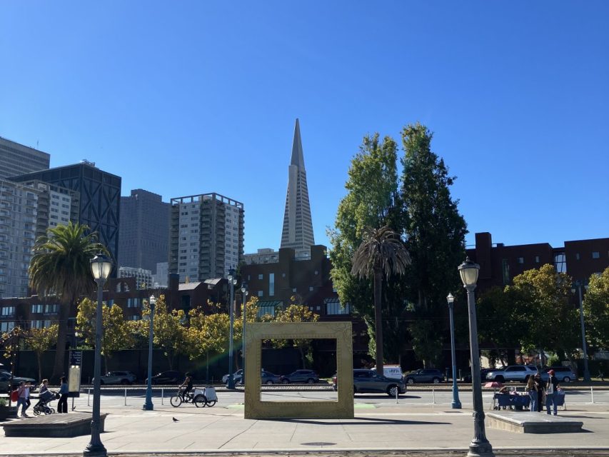 Cityscape with the Transamerica Pyramid in the background, trees and buildings in the foreground, people walking and a large rectangular sculpture in the center.