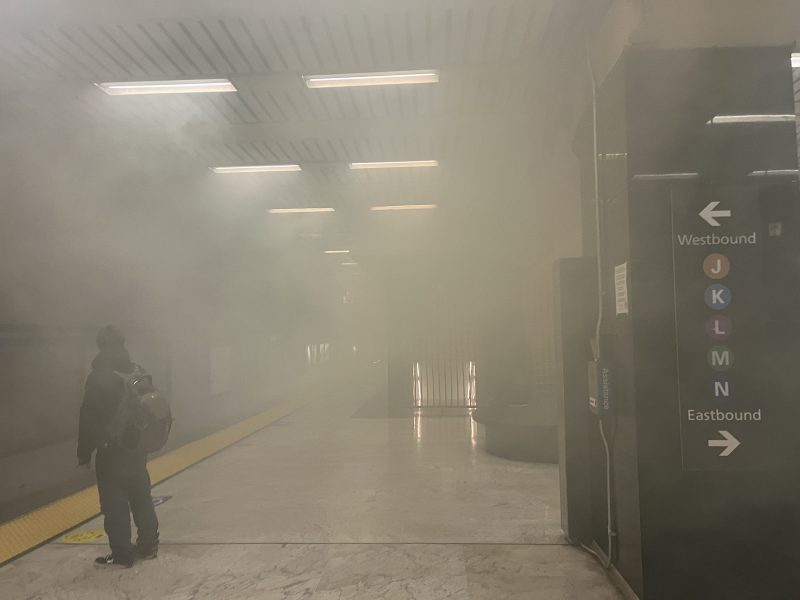 A subway platform filled with heavy smoke, with one person standing near the edge and directional signs for J, K, L, and M train lines visible.