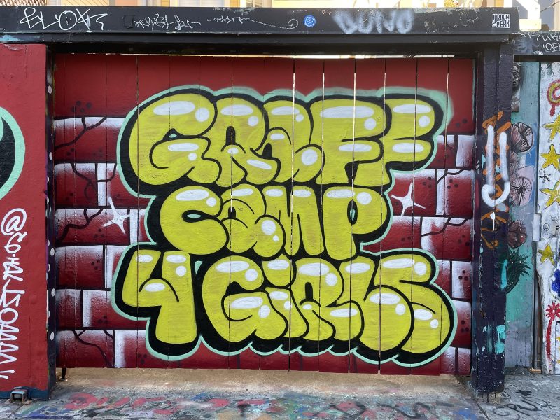 Yellow graffiti text reading "GRAFF CAMP 4 GIRLS" on a red and white mural wall, surrounded by other street art and graffiti tags.