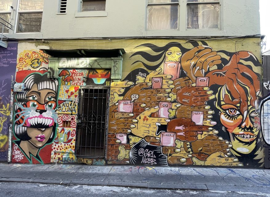 Colorful street art on a building wall features abstract faces, hands holding pink boxes, and vibrant patterns surrounding a gated doorway.