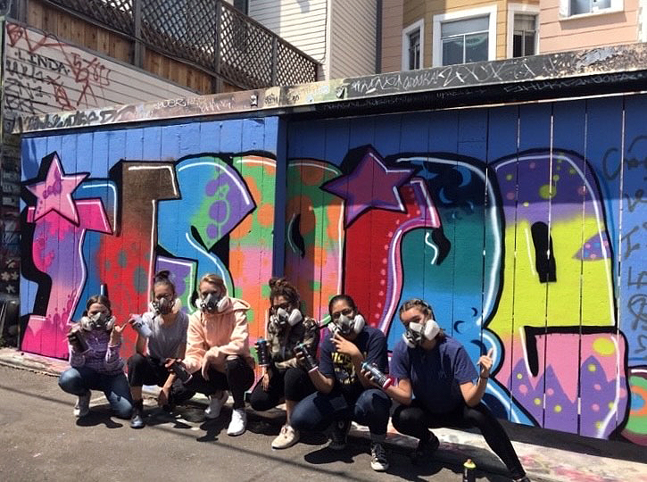 A group of six people wearing masks crouch in front of a colorful graffiti mural on a garage door in an urban alley.