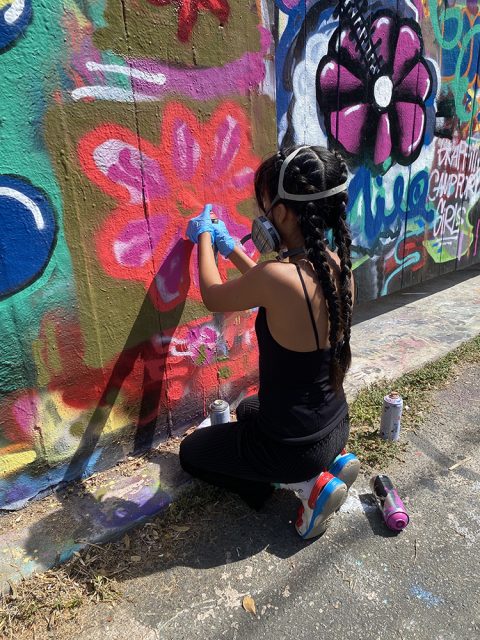 Person with long braided hair, wearing a mask and gloves, spray painting a red flower on a colorful graffiti-covered wall outdoors.