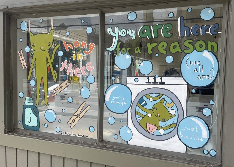 Storefront window art showing a green cat hanging from a clothesline, a cat in a washing machine, and motivational text like "hang in there" and "you are here for a reason.