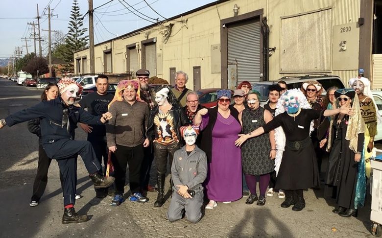 A group of people, some wearing colorful face paint and costumes, pose together on an industrial street in front of warehouse buildings.