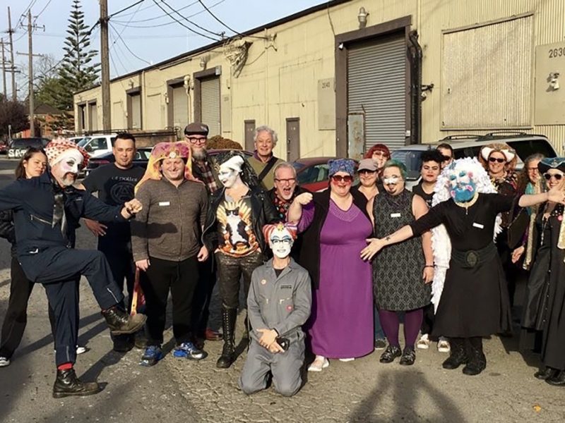 A group of people, some wearing colorful face paint and costumes, pose together on an industrial street in front of warehouse buildings.