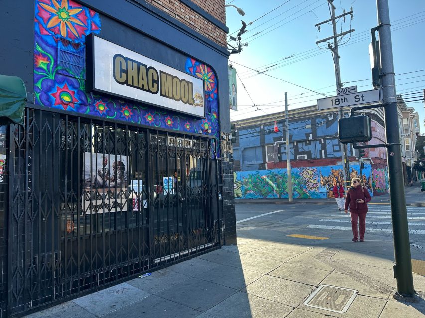Street view of Chac Mool storefront with colorful mural, near 18th St and a crosswalk; a person walks on the sidewalk under clear daylight.