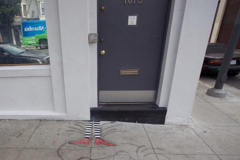 A gray door with the number 1075 above it; below the door, a mural depicts striped legs and red shoes, reminiscent of "The Wizard of Oz," painted on the sidewalk.