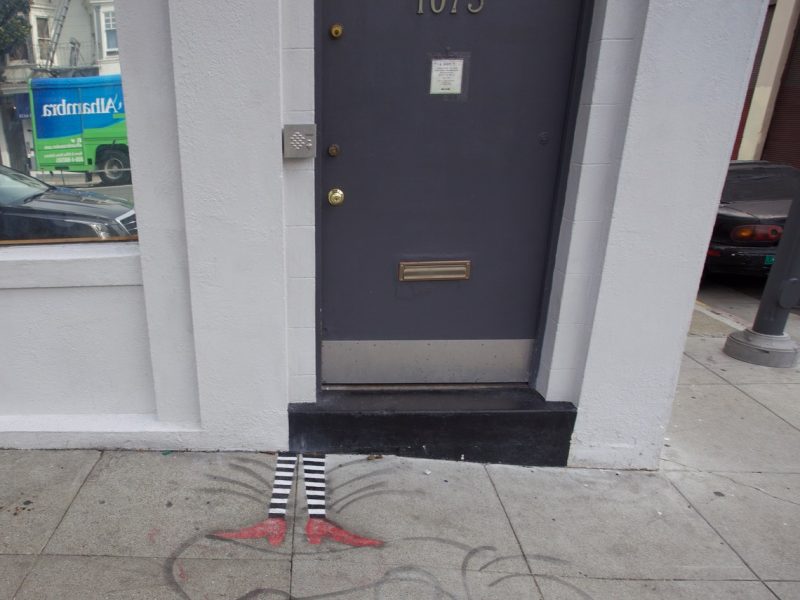 A gray door with the number 1075 above it; below the door, a mural depicts striped legs and red shoes, reminiscent of "The Wizard of Oz," painted on the sidewalk.
