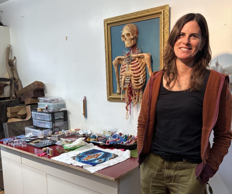 A woman stands next to a table with art supplies and fabric, with a framed anatomical skeleton model on the wall behind her.
