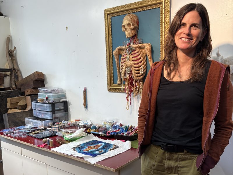 A woman stands next to a table with art supplies and fabric, with a framed anatomical skeleton model on the wall behind her.