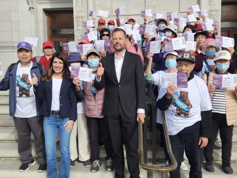A group of people stand on building steps, holding flyers and giving thumbs up; two people in front wear business attire, others wear casual clothing and hats.