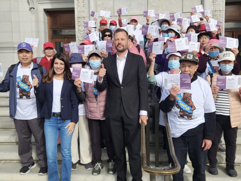 A group of people stand on building steps, holding flyers and giving thumbs up; two people in front wear business attire, others wear casual clothing and hats.