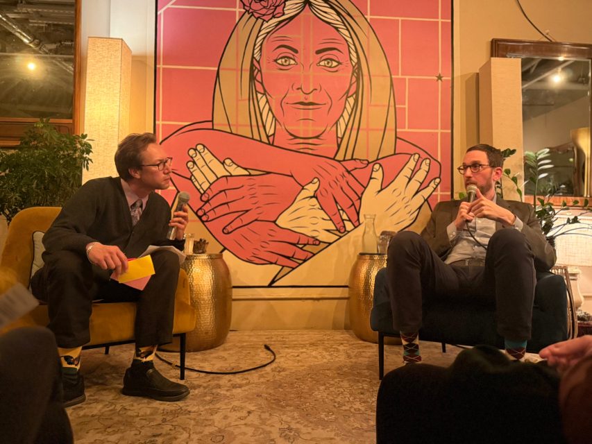 Two men sit and talk in armchairs in front of a large artwork featuring a woman with crossed arms. One man holds notes; both appear to be engaged in conversation.