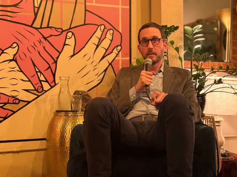 A man in glasses and a suit jacket sits in a chair holding a microphone, with a mural of hands and plants in the background.