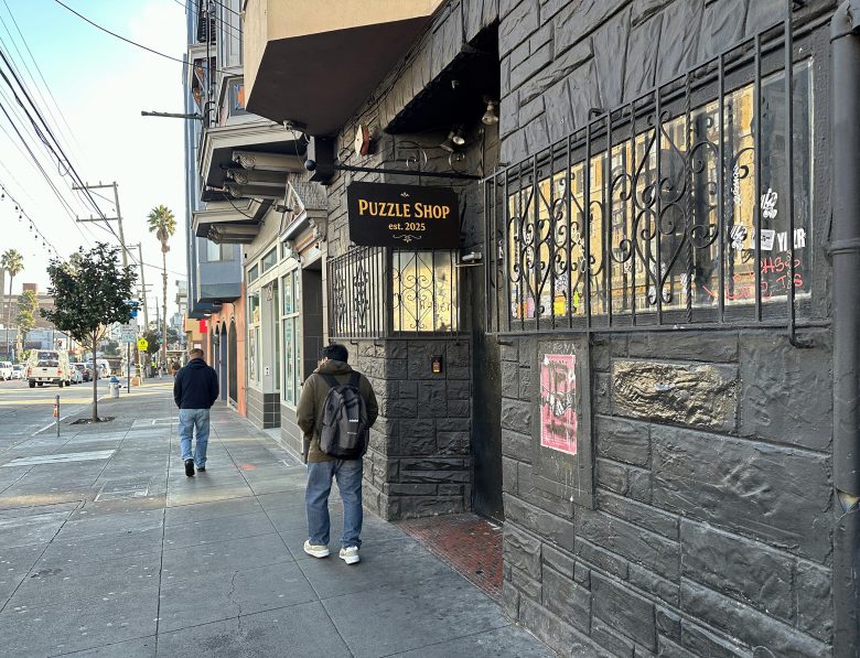 The exterior of a dark stone building with a sign reading "Puzzle Shop est. 2023." Two people walk on the sidewalk nearby. Urban street setting with some graffiti and posters.