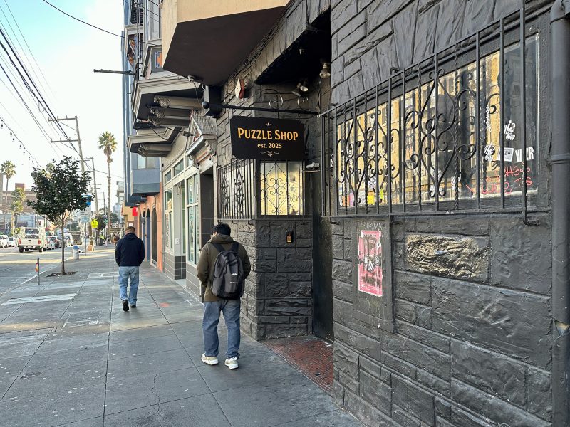 The exterior of a dark stone building with a sign reading "Puzzle Shop est. 2023." Two people walk on the sidewalk nearby. Urban street setting with some graffiti and posters.