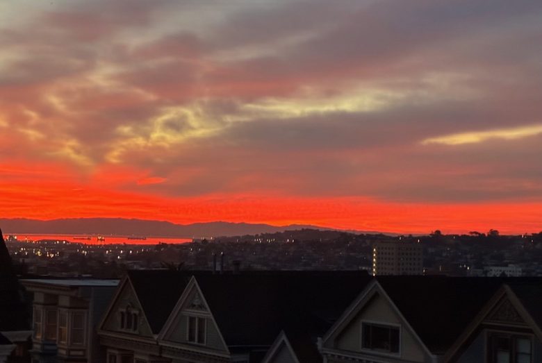 Fiery orange and red sunset sky over a cityscape with silhouetted rooftops and distant hills.