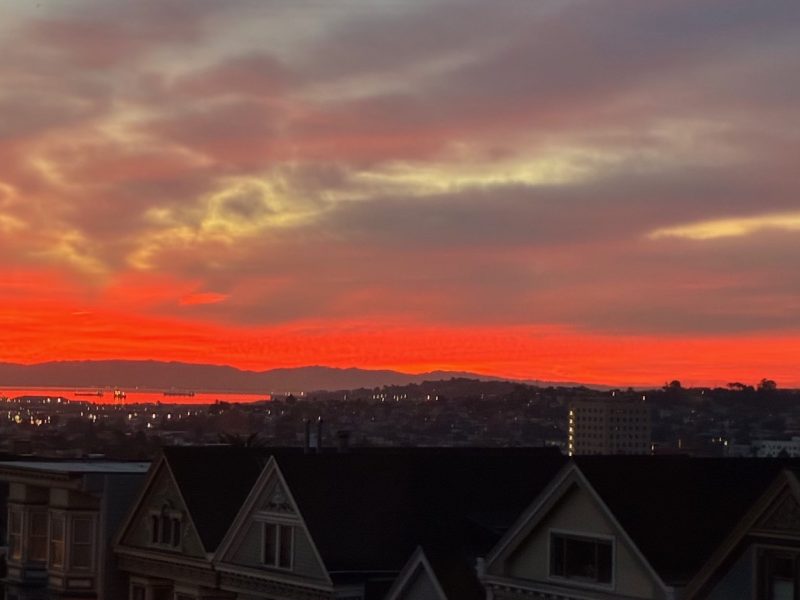 Fiery orange and red sunset sky over a cityscape with silhouetted rooftops and distant hills.