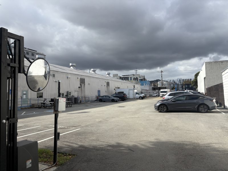 A parking lot next to a long white industrial building under a cloudy sky, with several cars parked and a security mirror in the foreground.