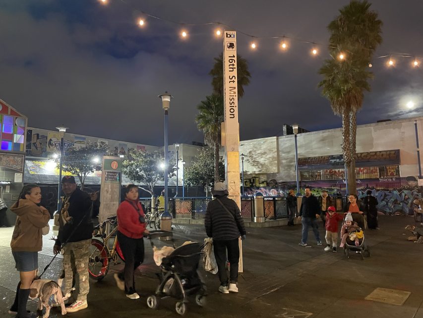 People walk and gather near the 16th St Mission BART station at night, with string lights overhead and graffiti-covered walls in the background.