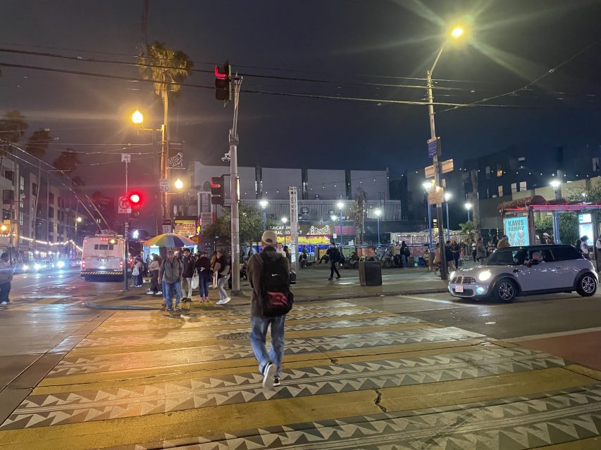 A busy city street at night with people crossing, cars stopped at a traffic light, and brightly lit street vendors and shops in the background.